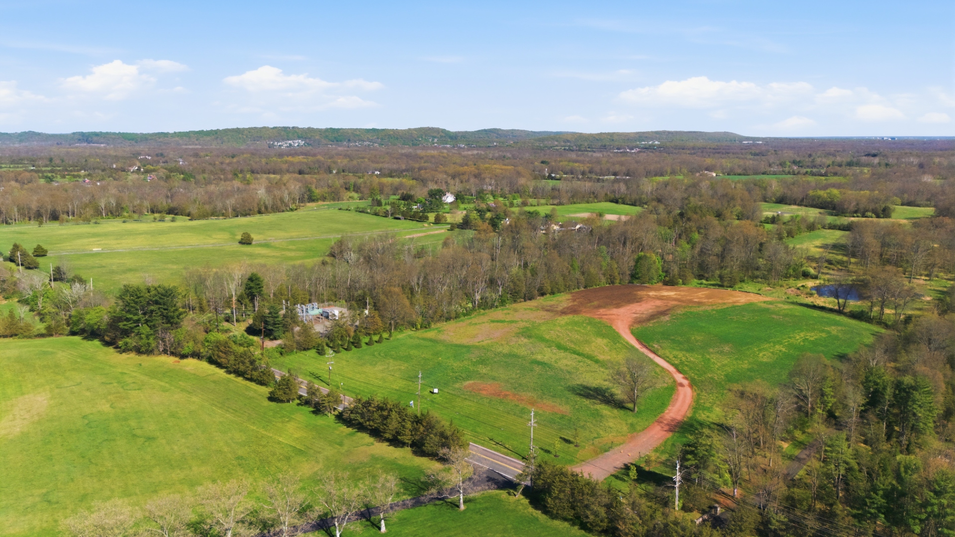 Sweeping aerial vista over the 122-acre Bedminster tract showing open meadow, a graded access road and the Somerset Hills on the horizon